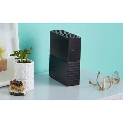External black hard drive next to a plant in a white pot and a pair of glasses on a desk in front of a turquoise wall.