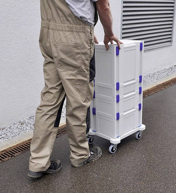 A man in workwear is pushing a stacked trolley with boxes along a building on a pavement.