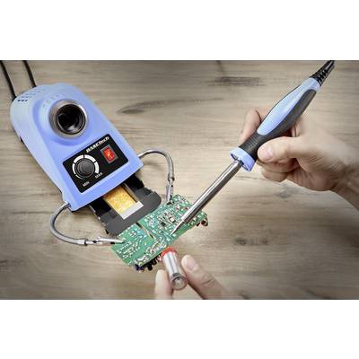 A soldering iron is used to solder an electronic circuit onto a green circuit board; a bright soldering station is in the background.