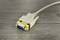 A grey VGA cable lies on a wooden table. It has blue pins and is wrapped with a yellow label that reads 'REMOVE BEFORE USE'.