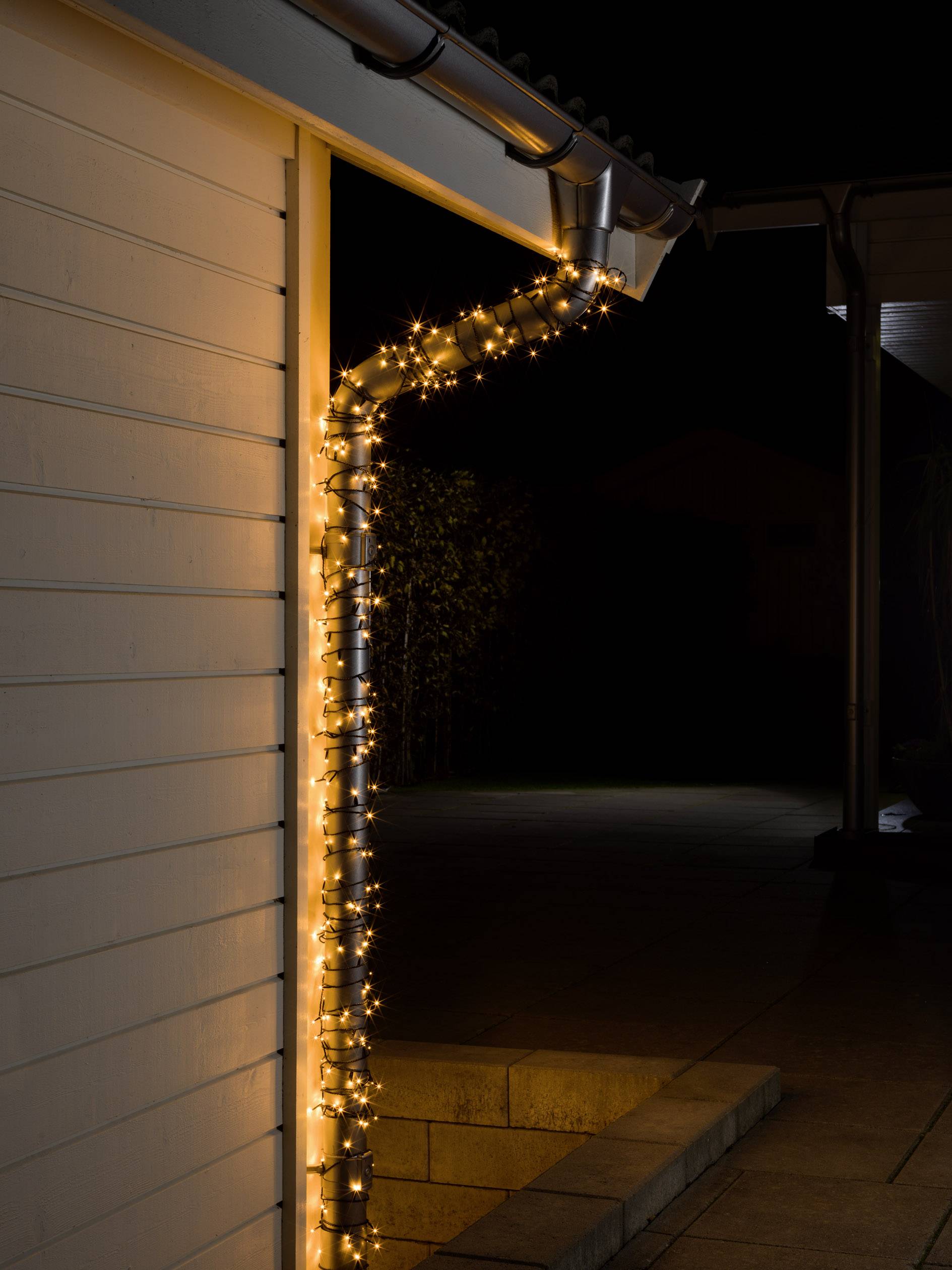 A string of fairy lights winds around a gutter on a house at night, providing festive lighting. Background dark.