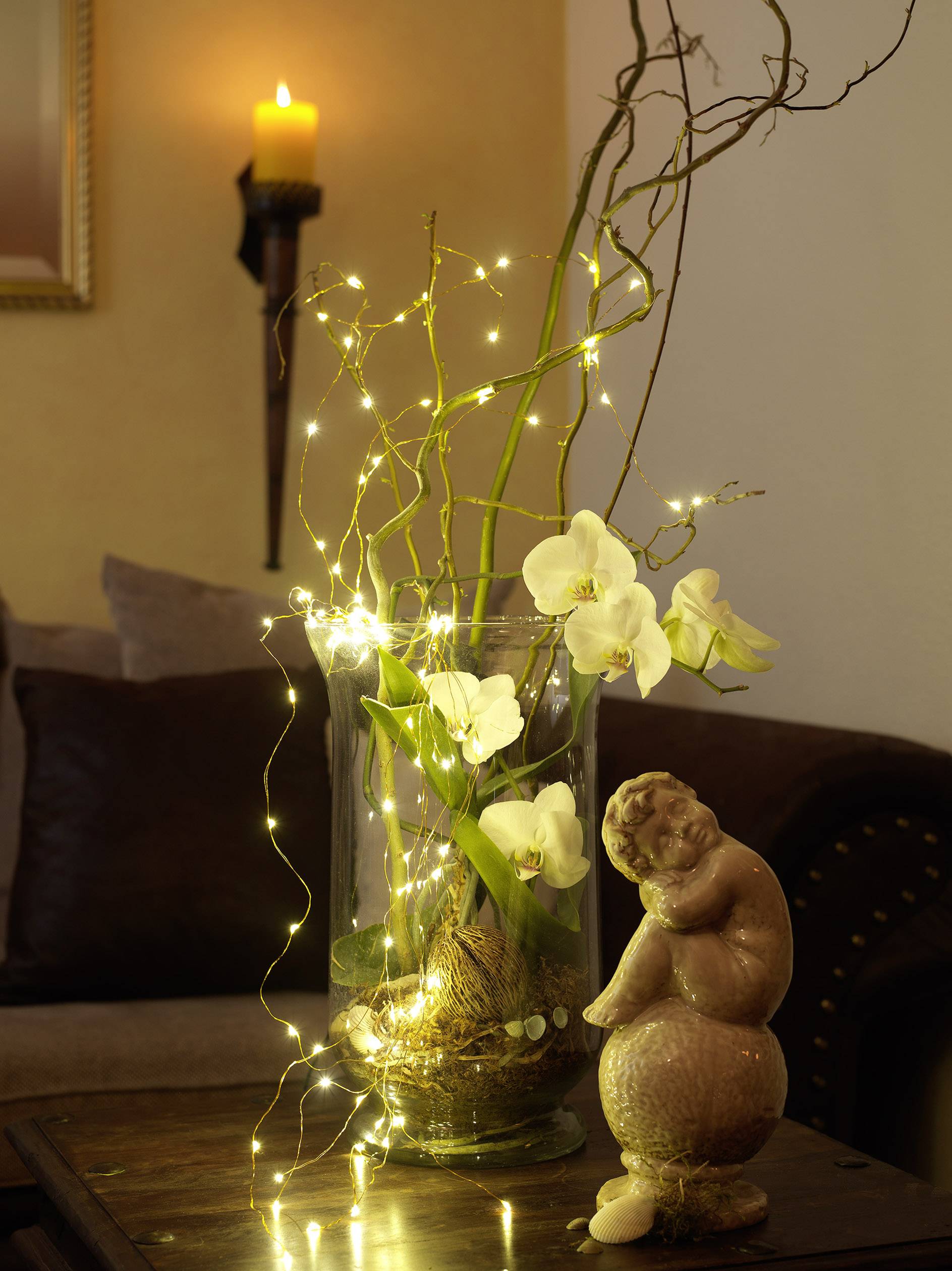 A vase with white orchids and twinkling fairy lights sits on a table, beside a statue. In the background, a candle is burning.