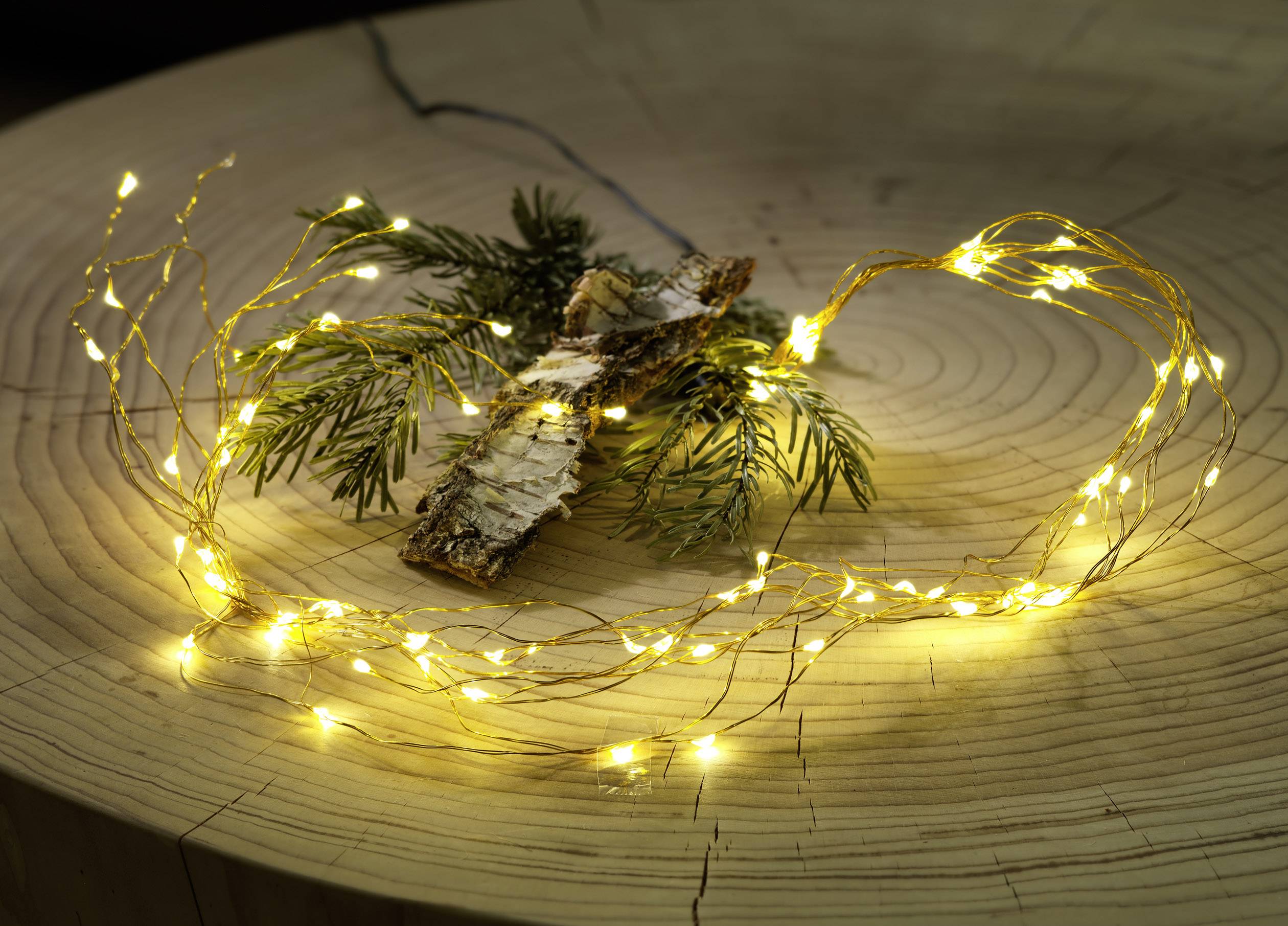 A small yellow fairy light string and pine branch with bark arranged on a wooden table, conveying a cosy, festive atmosphere.