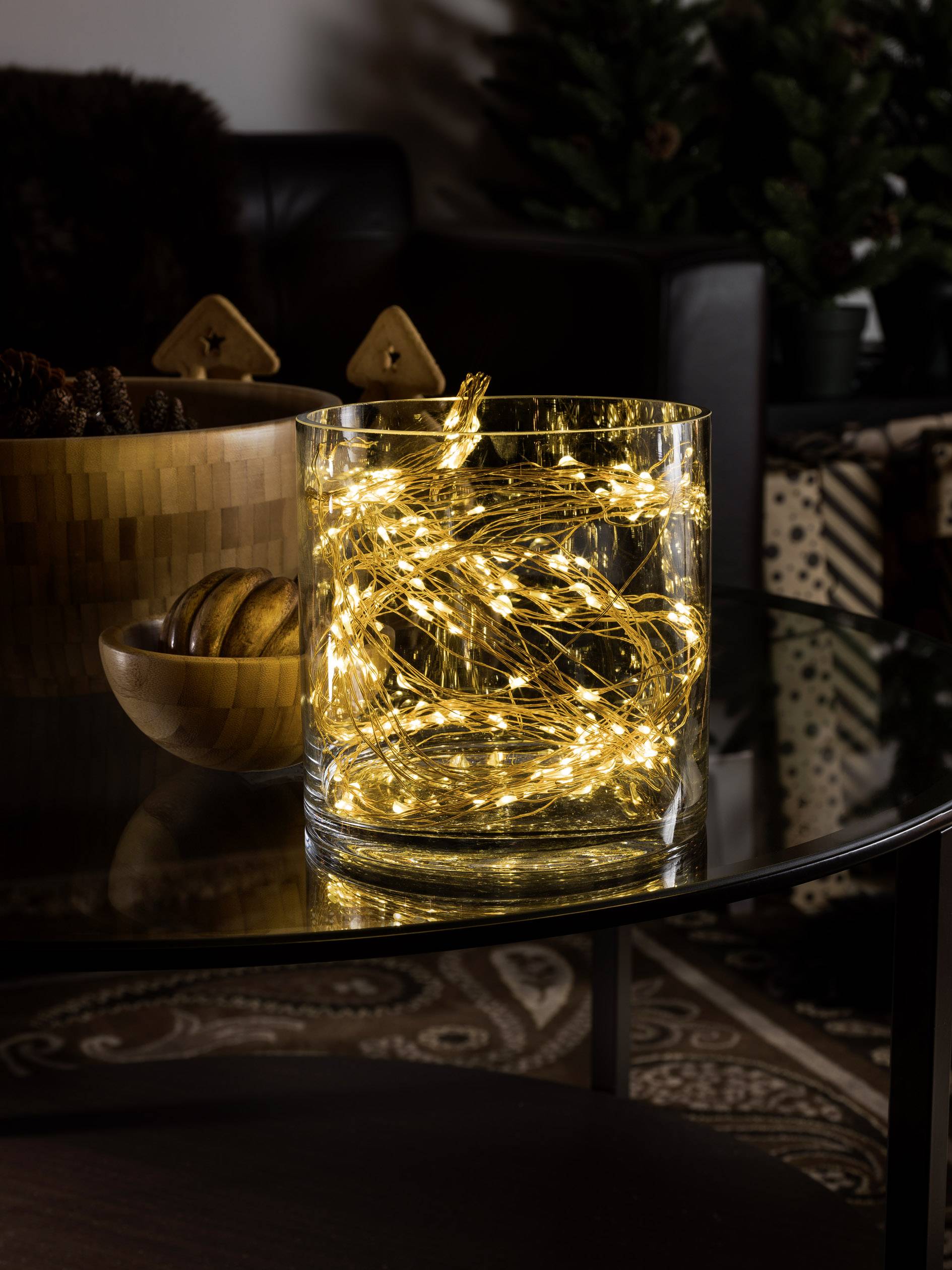 A glass vessel on a table contains illuminated fairy lights. In the background, wooden arches and a plant are visible.