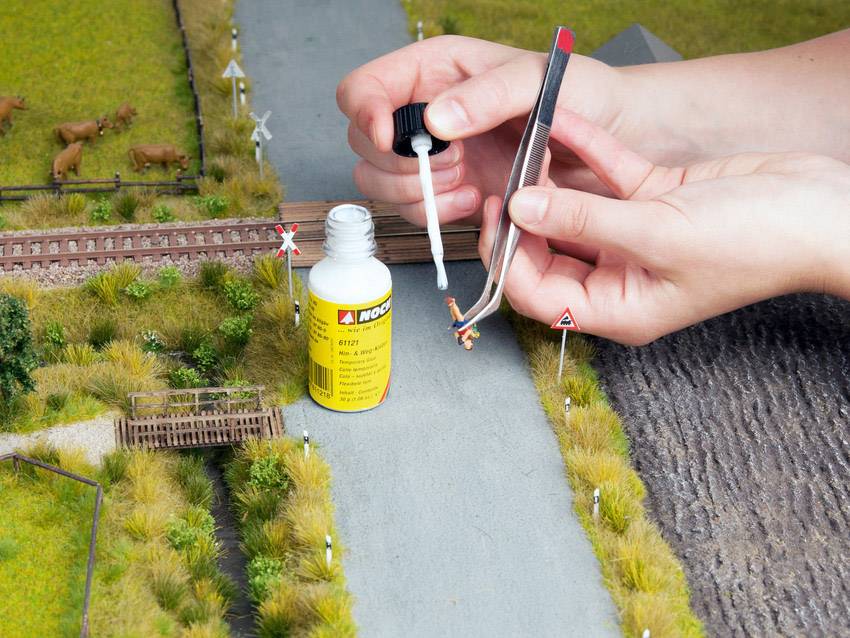 A person is sticking a small model with glue and tweezers onto a model railway landscape with tracks and grass.