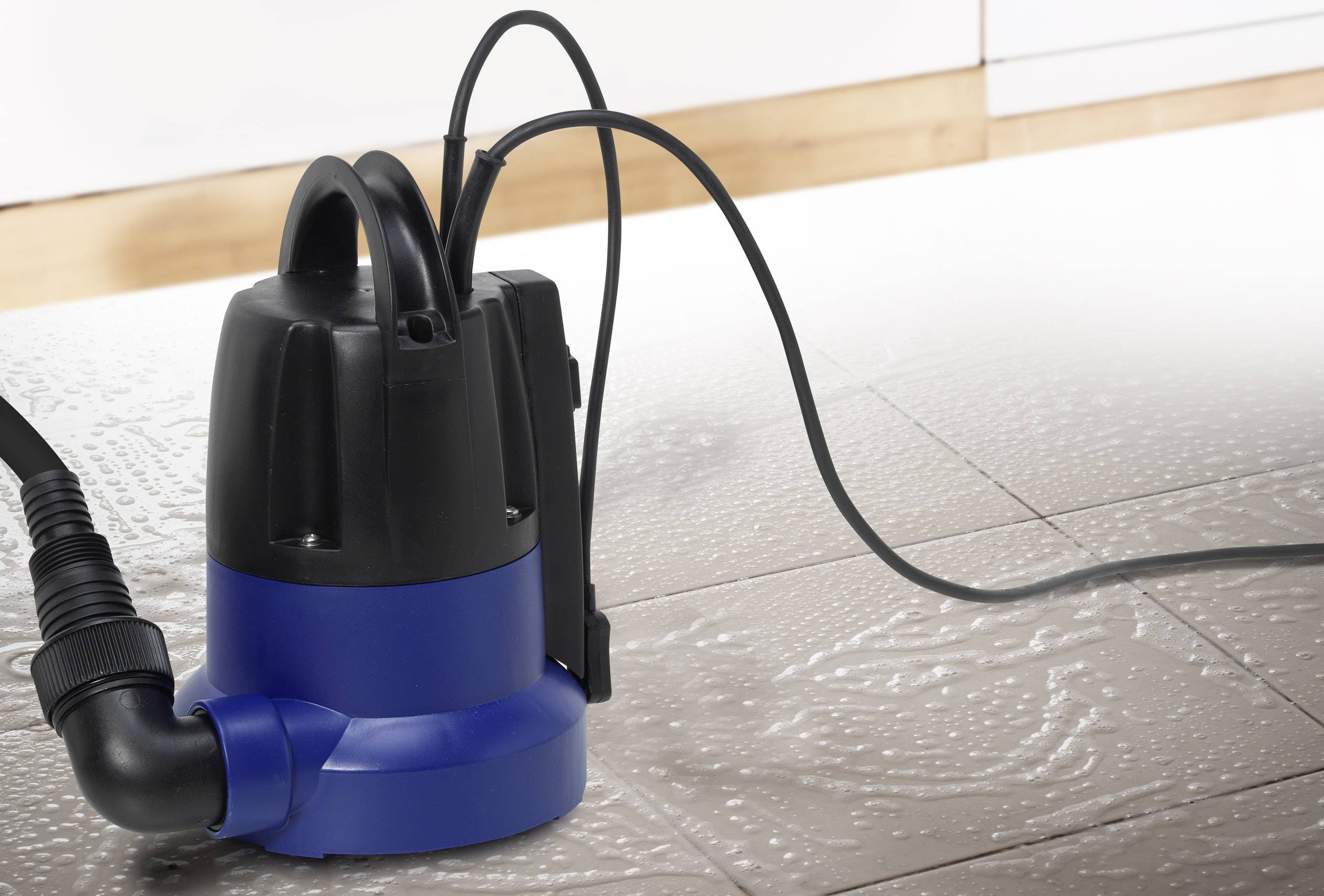 A submersible pump on a flooded tiled floor, ready to pump out water. Background shows a wooden plinth against the wall.