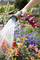A hand holds a garden hose and waters colourful flowers in the garden. The flowers are red, blue and pink and are blooming vibrantly.