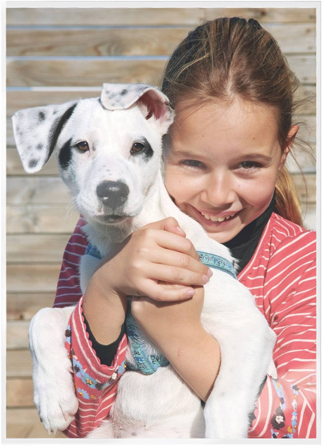 A smiling child hugs a white puppy with black spots. Both look happy. A wooden wall is in the background.