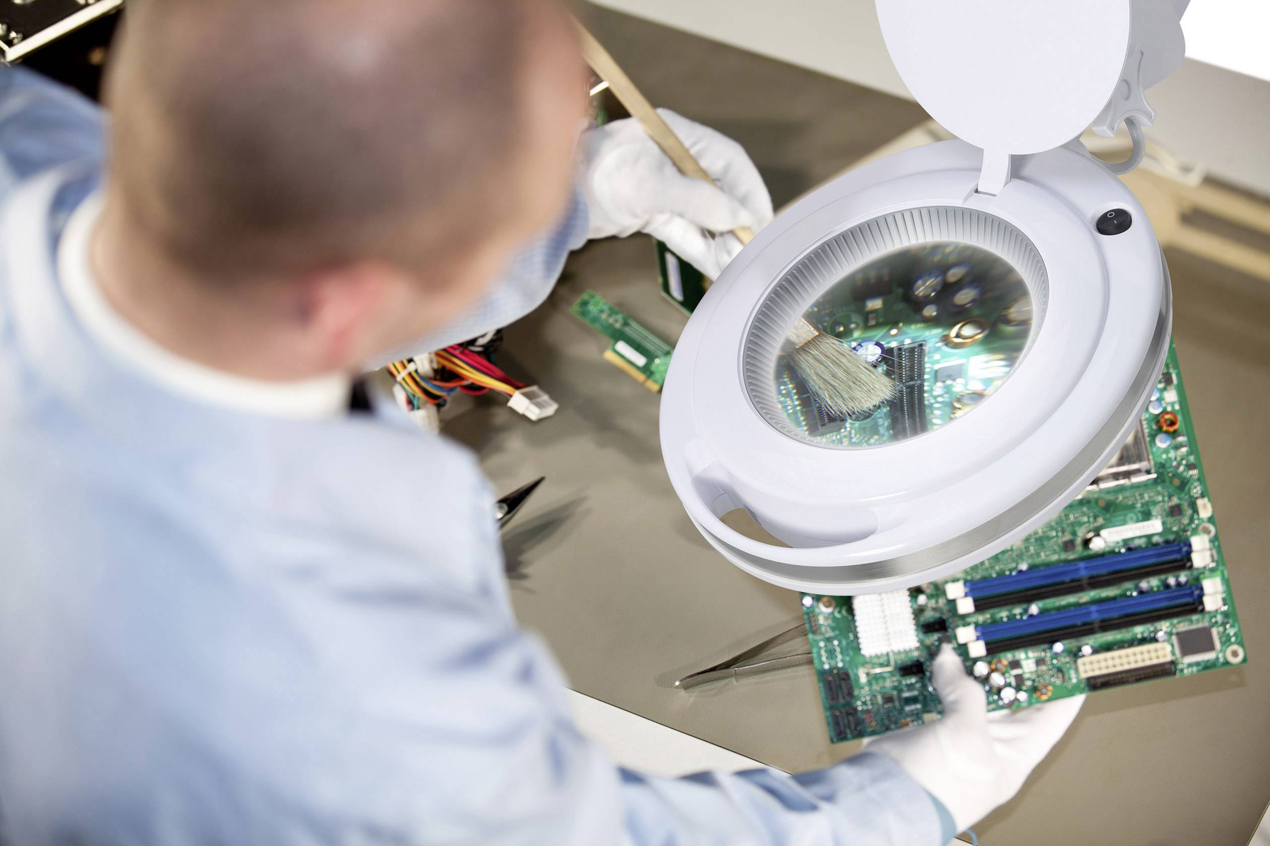 A technician cleans a circuit board under an illuminated magnifying glass with a brush.