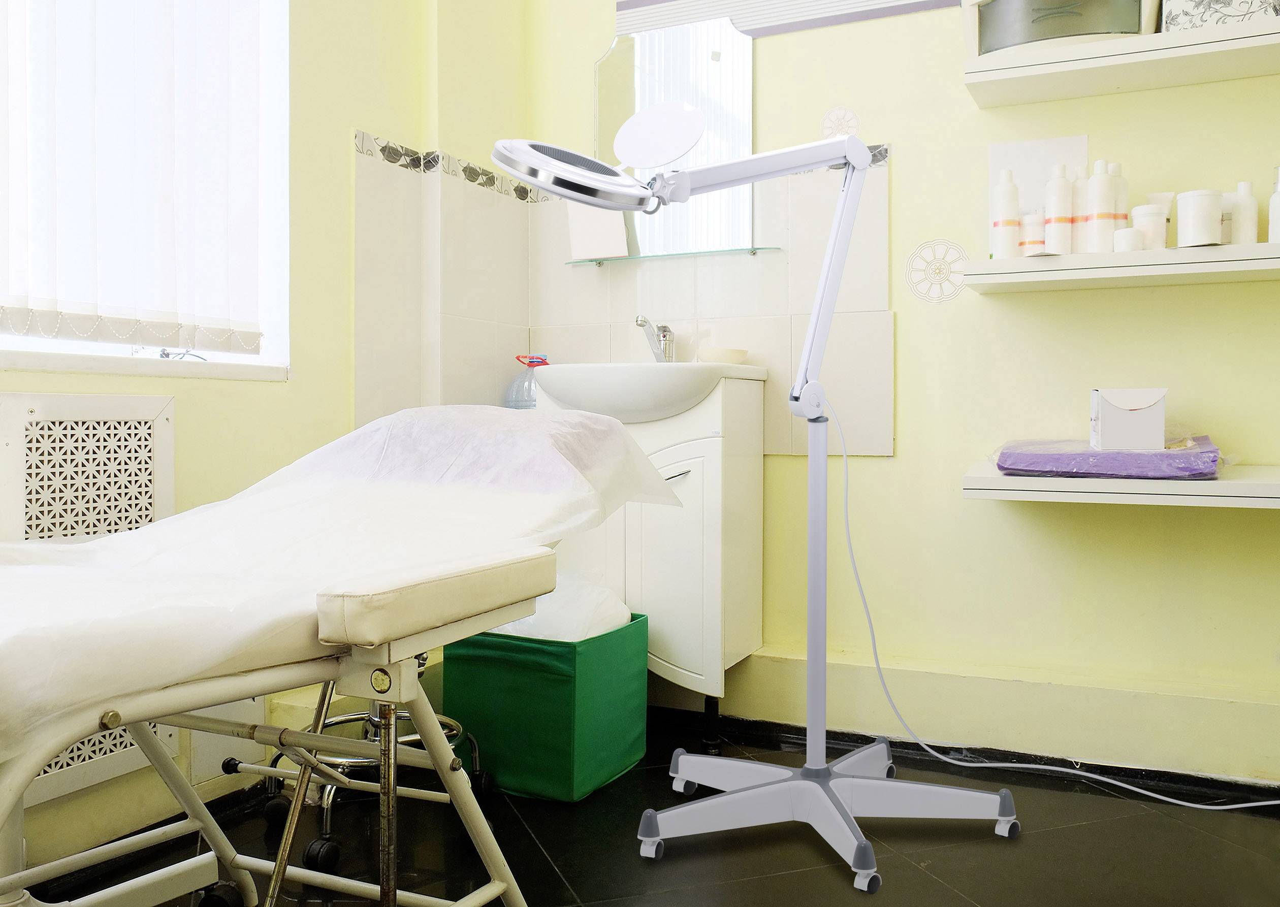 Beauty salon with a treatment bed, magnifying lamp and skincare products on shelves. Bright white walls and a washbasin in the background.