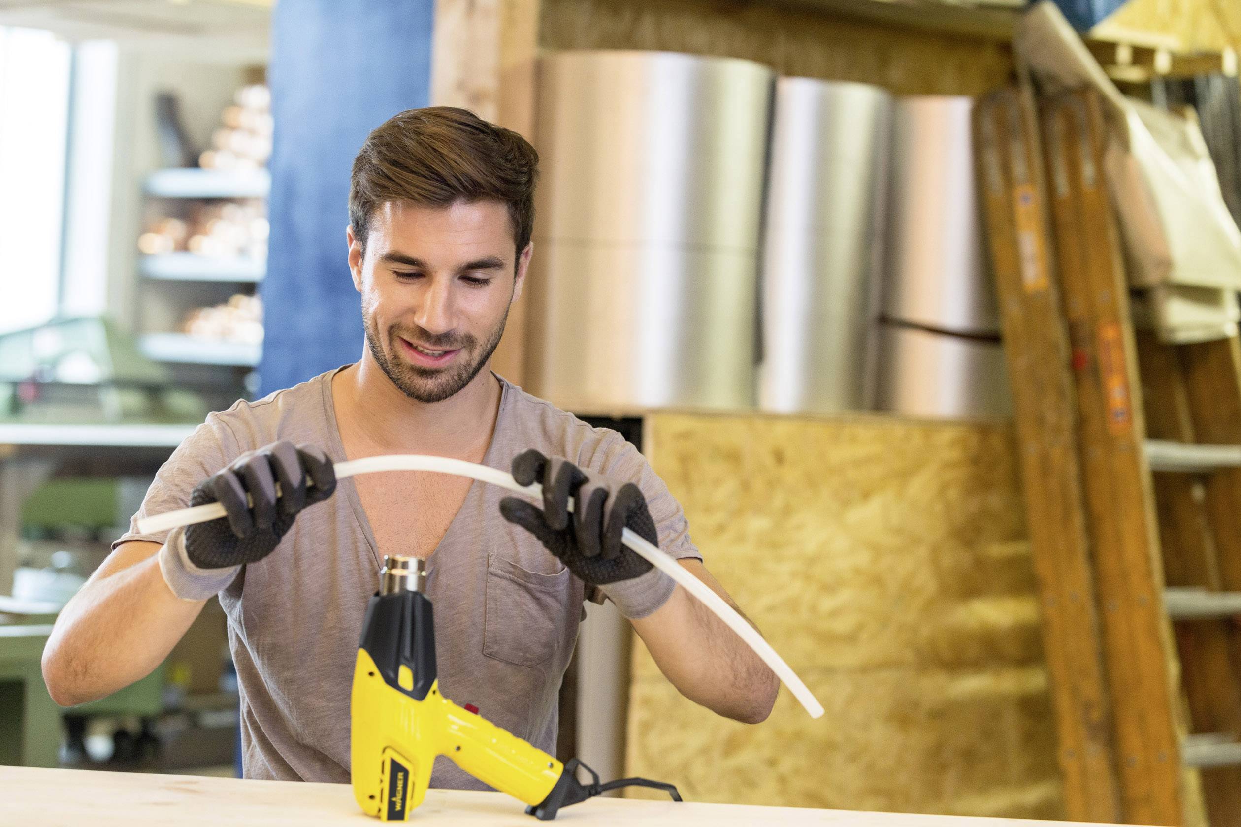 A man wearing gloves is bending a plastic pipe using a hot air blower in a workshop.