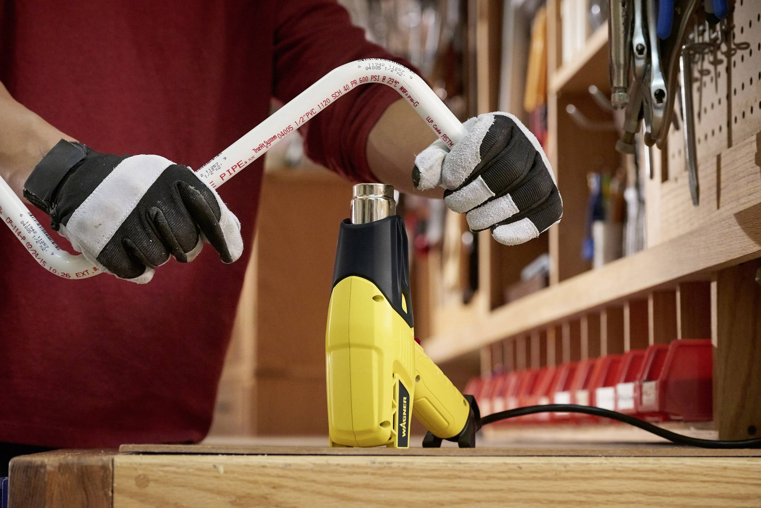 A person is bending a pipe using a yellow heat gun in a workshop. Tools are hanging on a wall to the left.
