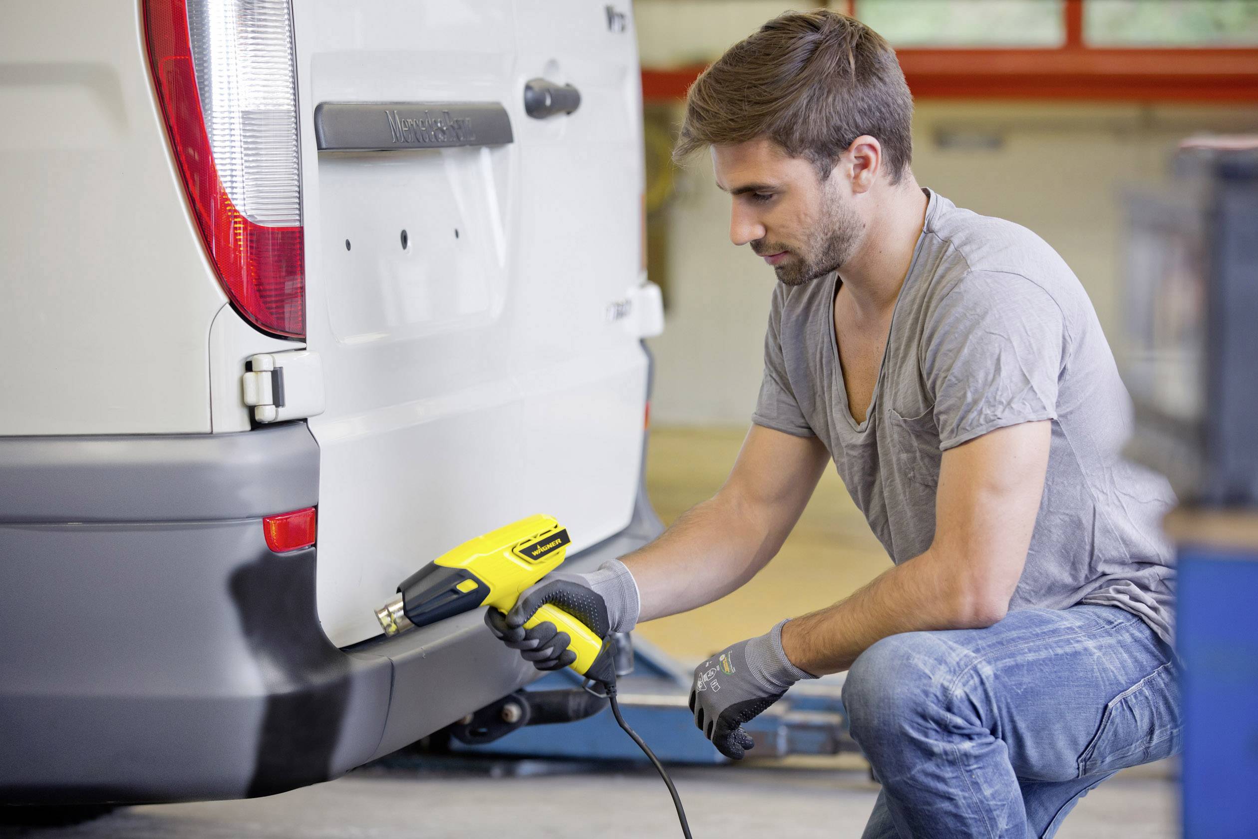 A man in a grey t-shirt is using a hot air blower to repair the bumper of a white van.