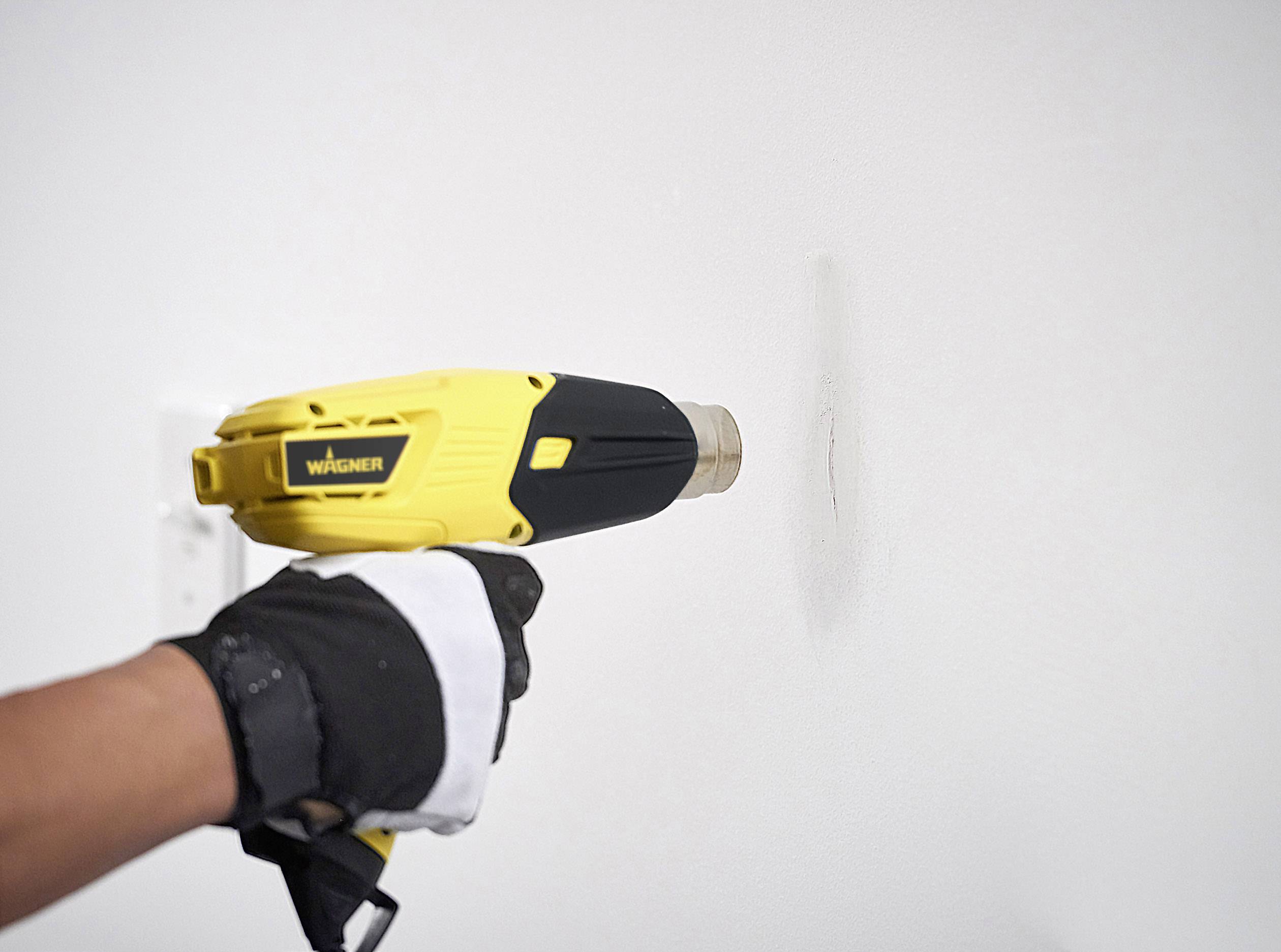 A person is holding a hot air blower against a wall to soften the paint. A glove protects the hand from heat.
