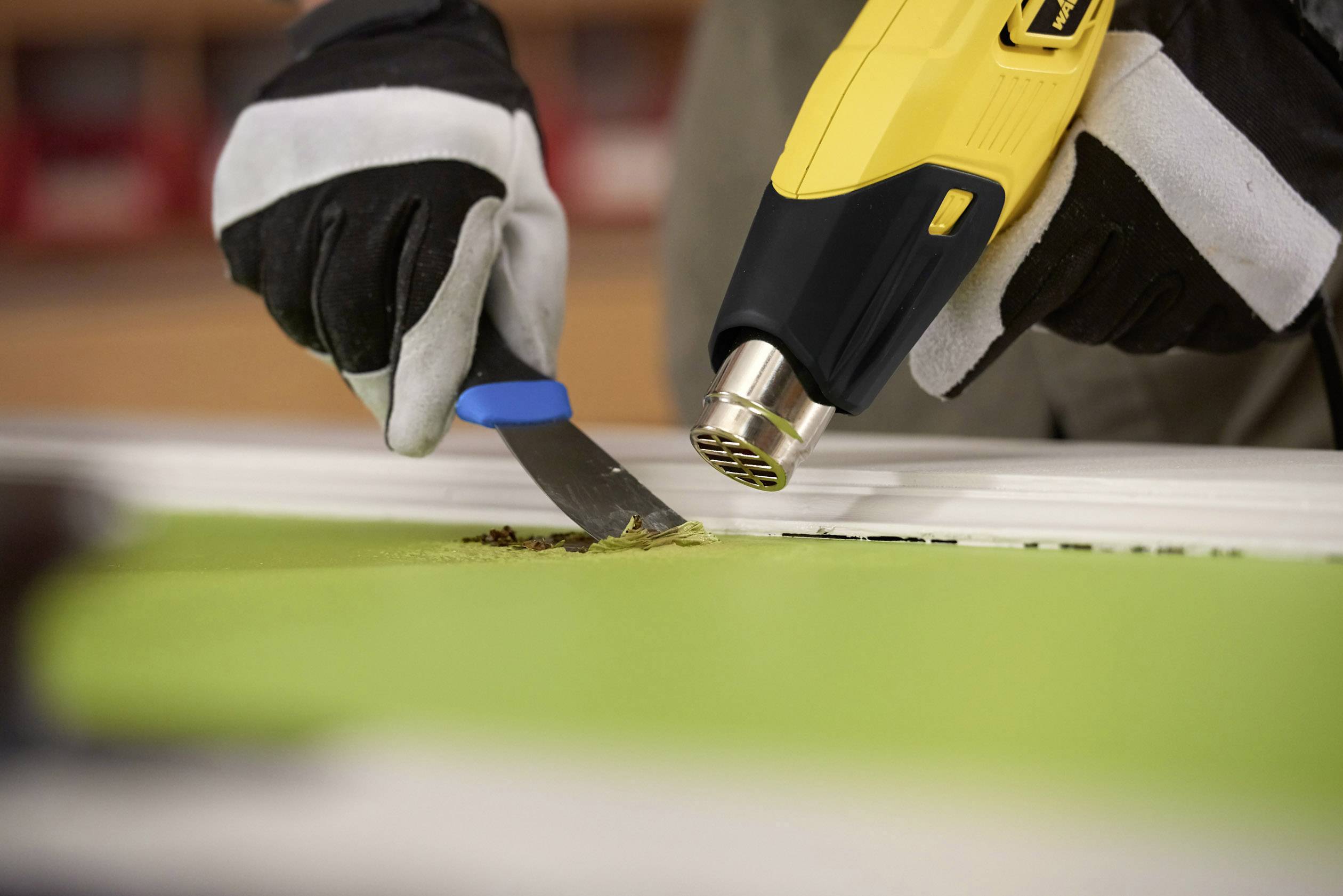 'Two gloved hands remove paint from a green surface using a yellow hot air dryer and a scraper.'