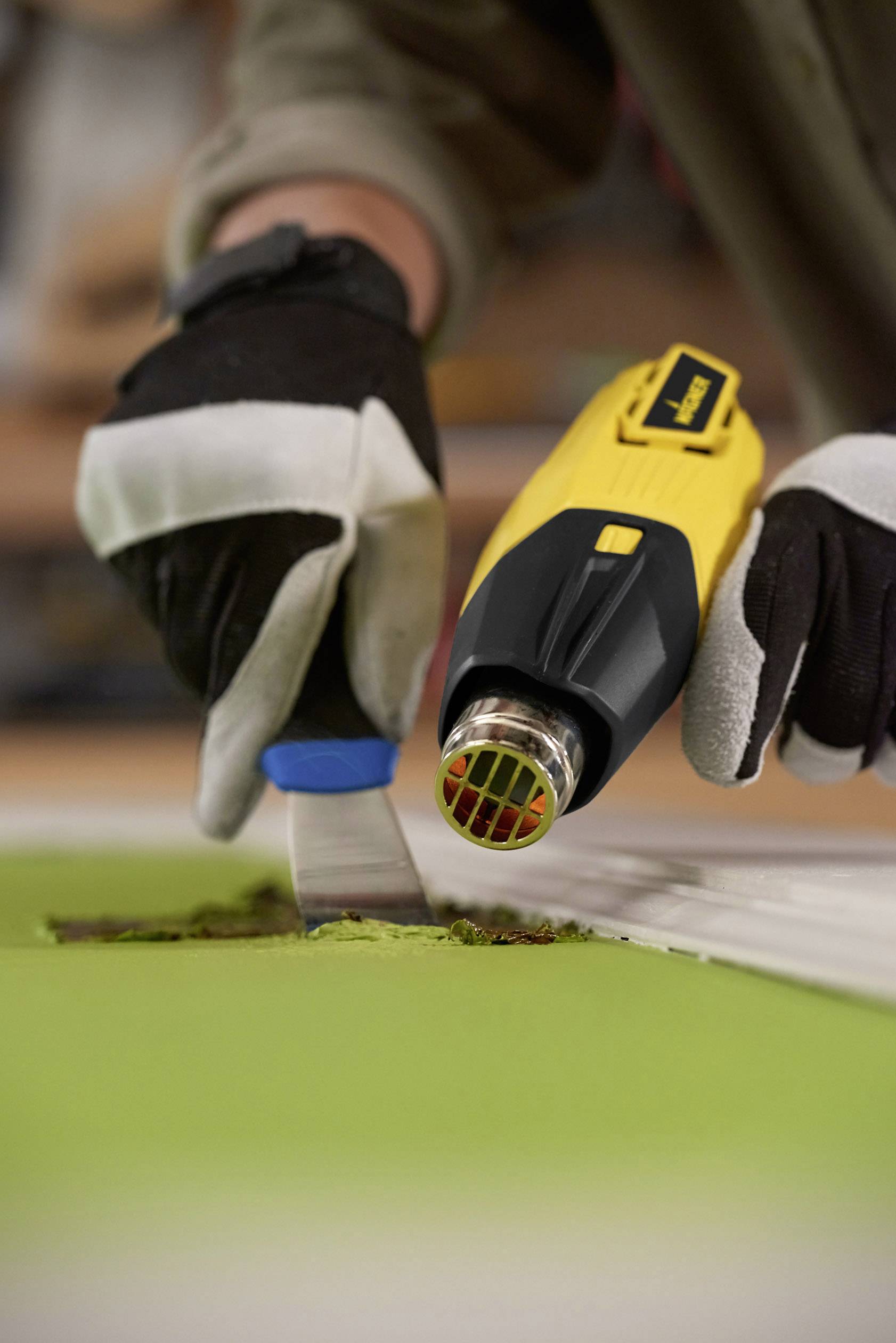 A person is removing paint from a green surface using a yellow heat gun and a scraper. They are wearing work gloves.