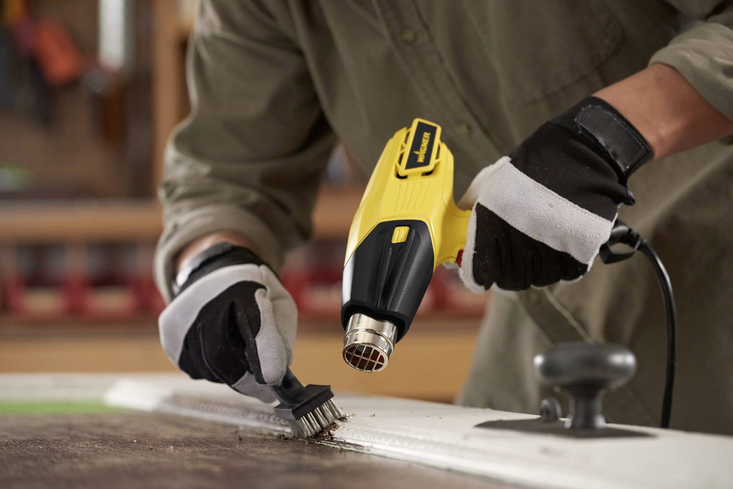 A person is removing old paint from a wooden surface using a yellow hot air gun and a brush, wearing work gloves.