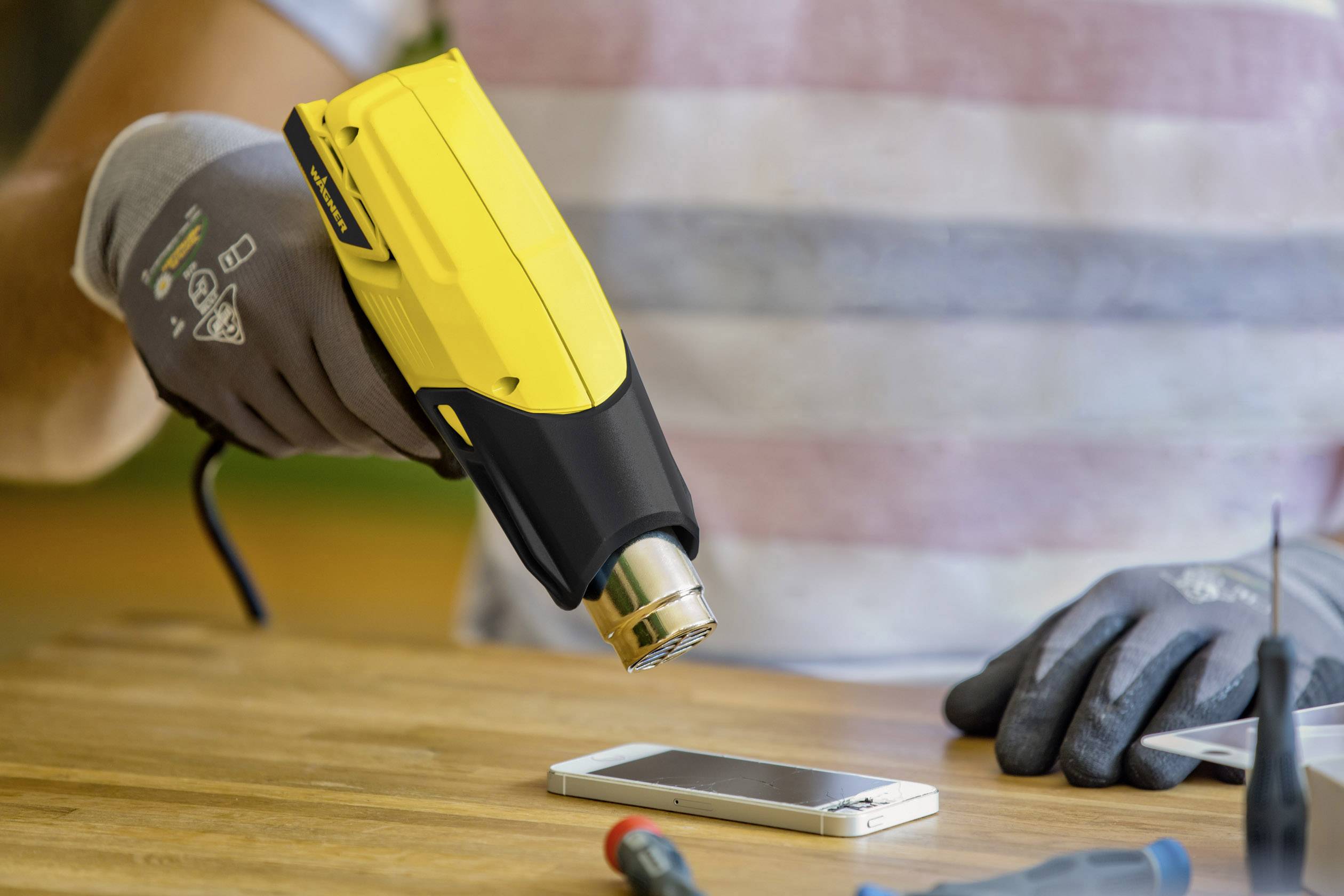 A person is heating a smartphone on a wooden table with a yellow hot air blower, presumably for repair work.
