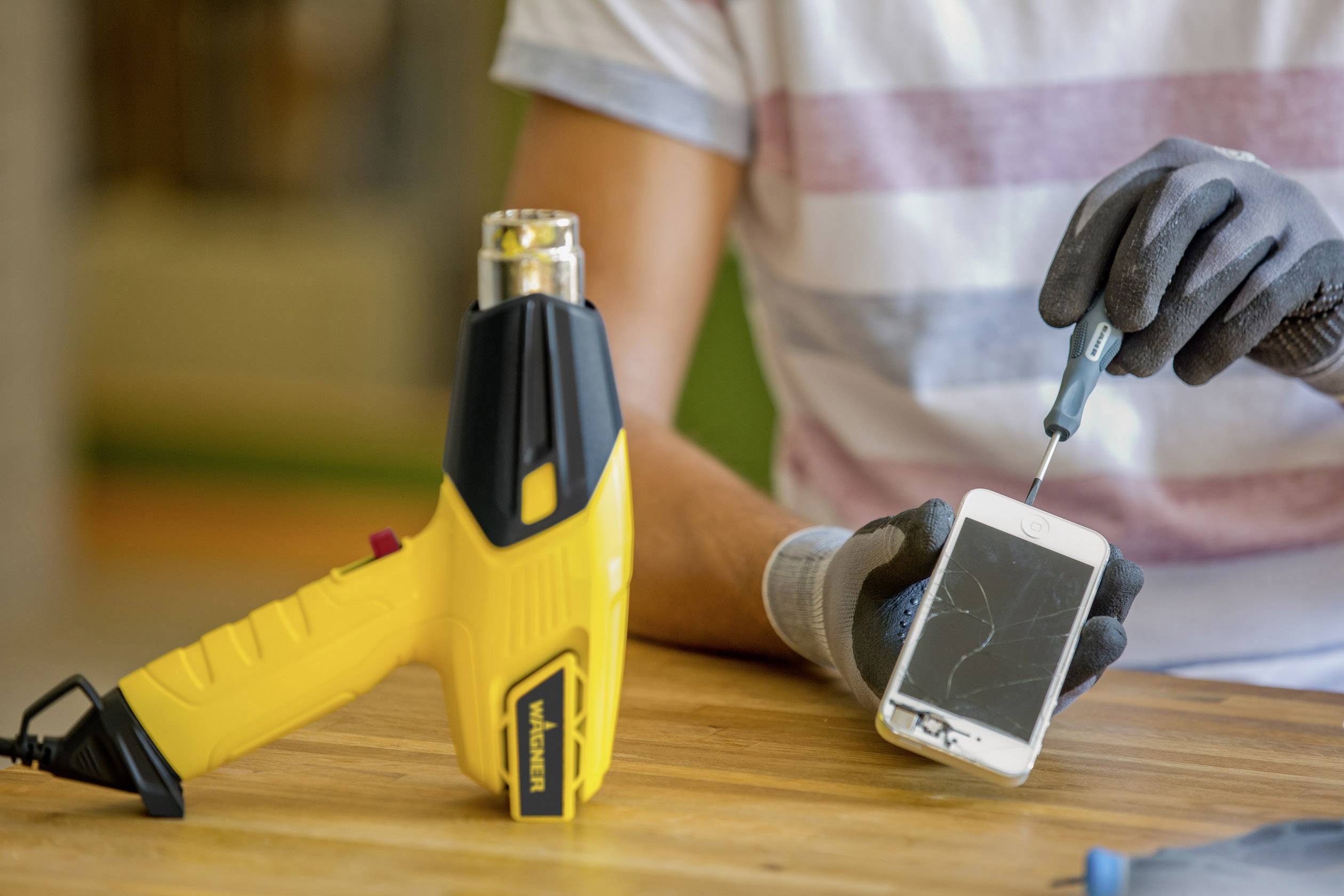 A person is repairing a smartphone screen with a hot air blower and tools. The screen is cracked. Hands are wearing protective gloves.