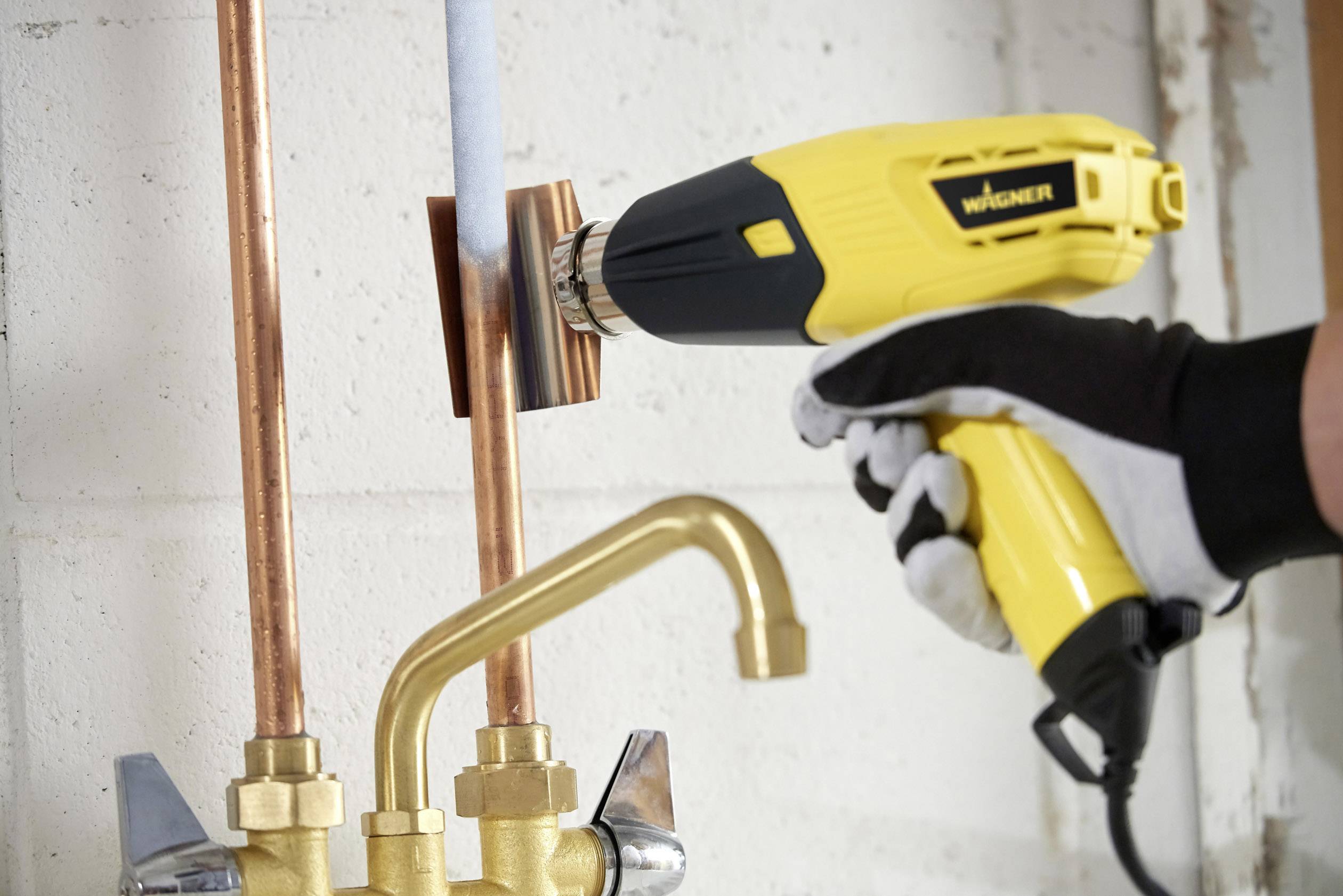 A person is heating a copper pipe connection with a yellow hot air blower next to other pipes and a tap.