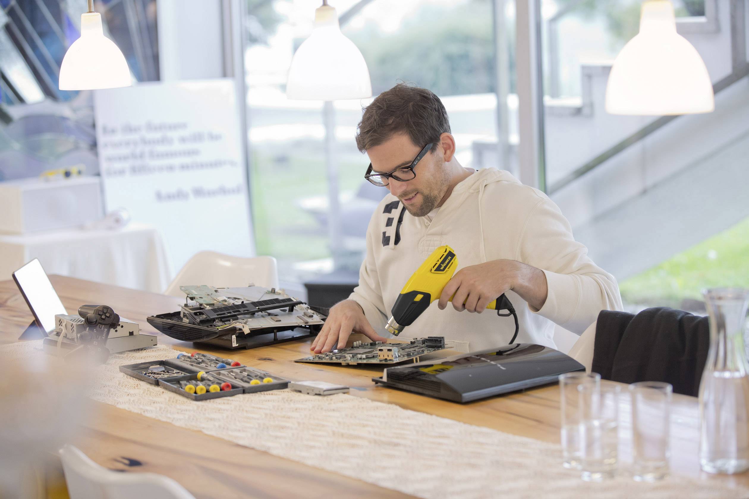 A person is sitting at a table and repairing electronic devices with a soldering iron. Various tools and components are scattered around.