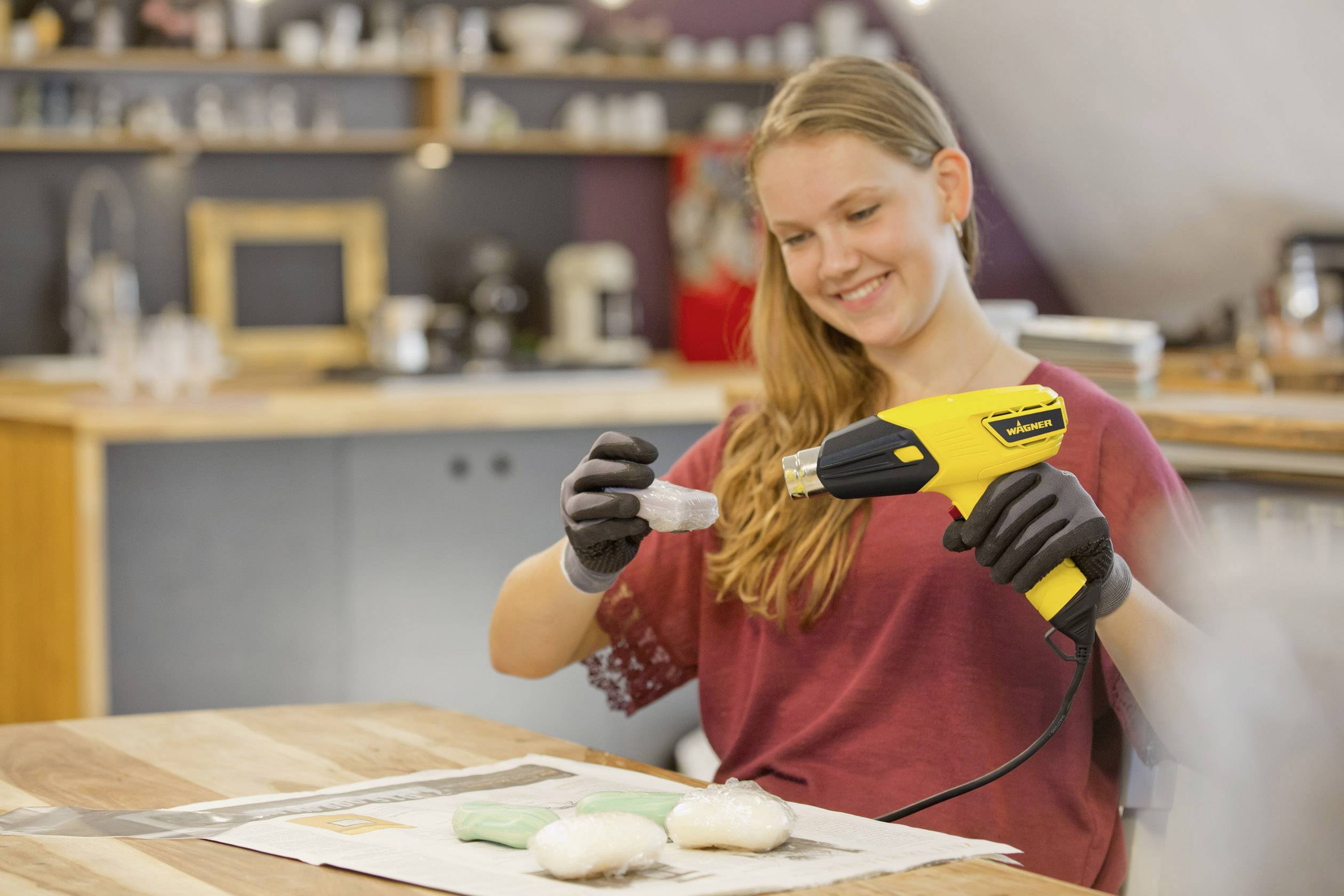 A woman in a workshop is using a heat gun to shrink plastic film around a small object. She is wearing gloves.