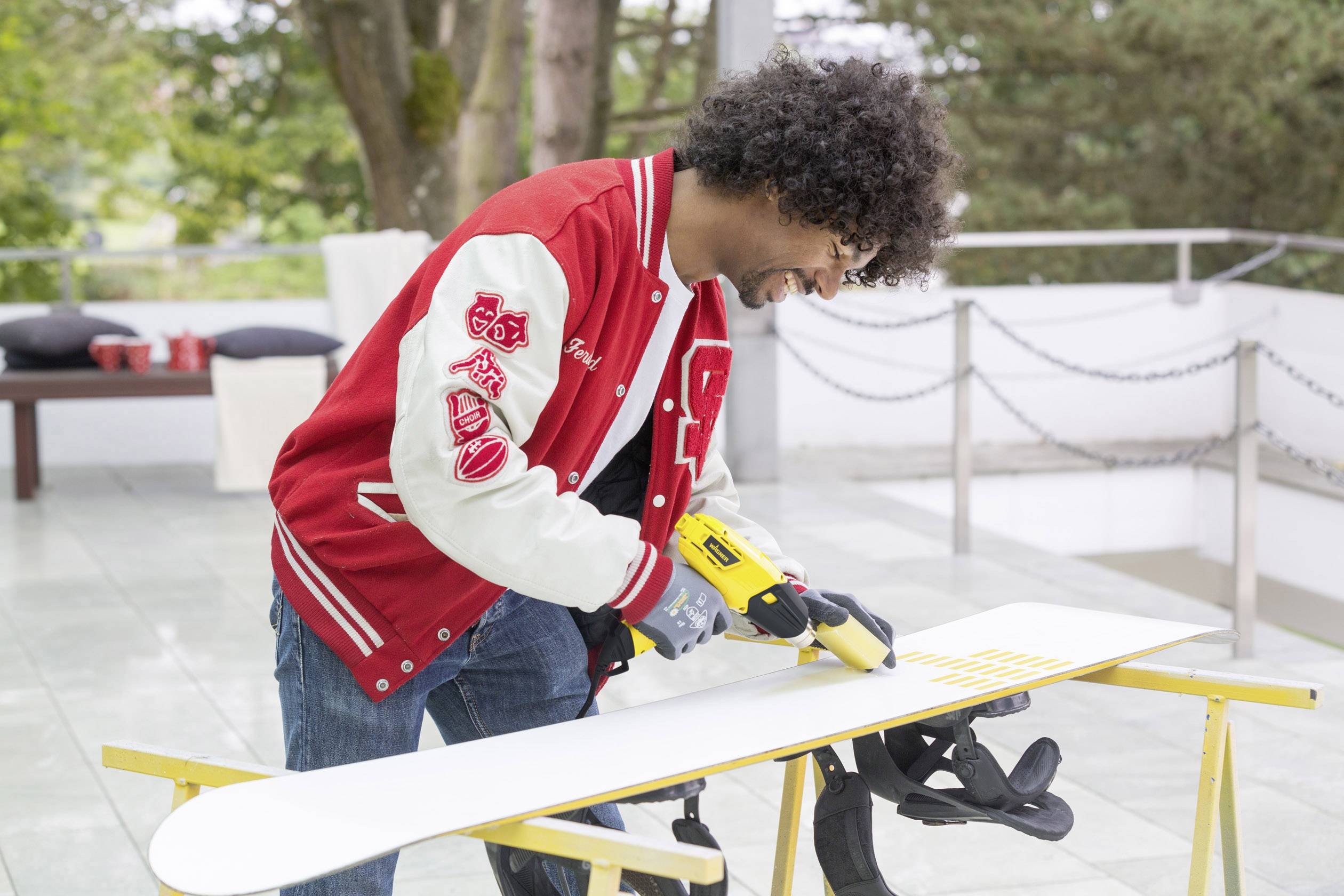 A person is waxing a snowboard on an outdoor table. They are wearing a red college jacket. Trees are visible in the background.