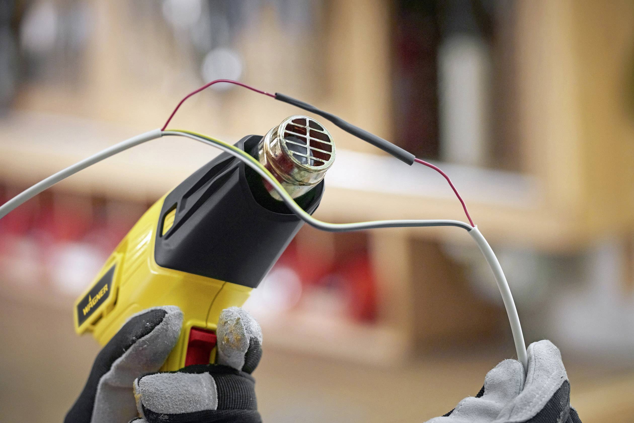 A person is working with a heating device that is heating a heat-shrink sleeve onto a cable. Background blurred with workshop environment.