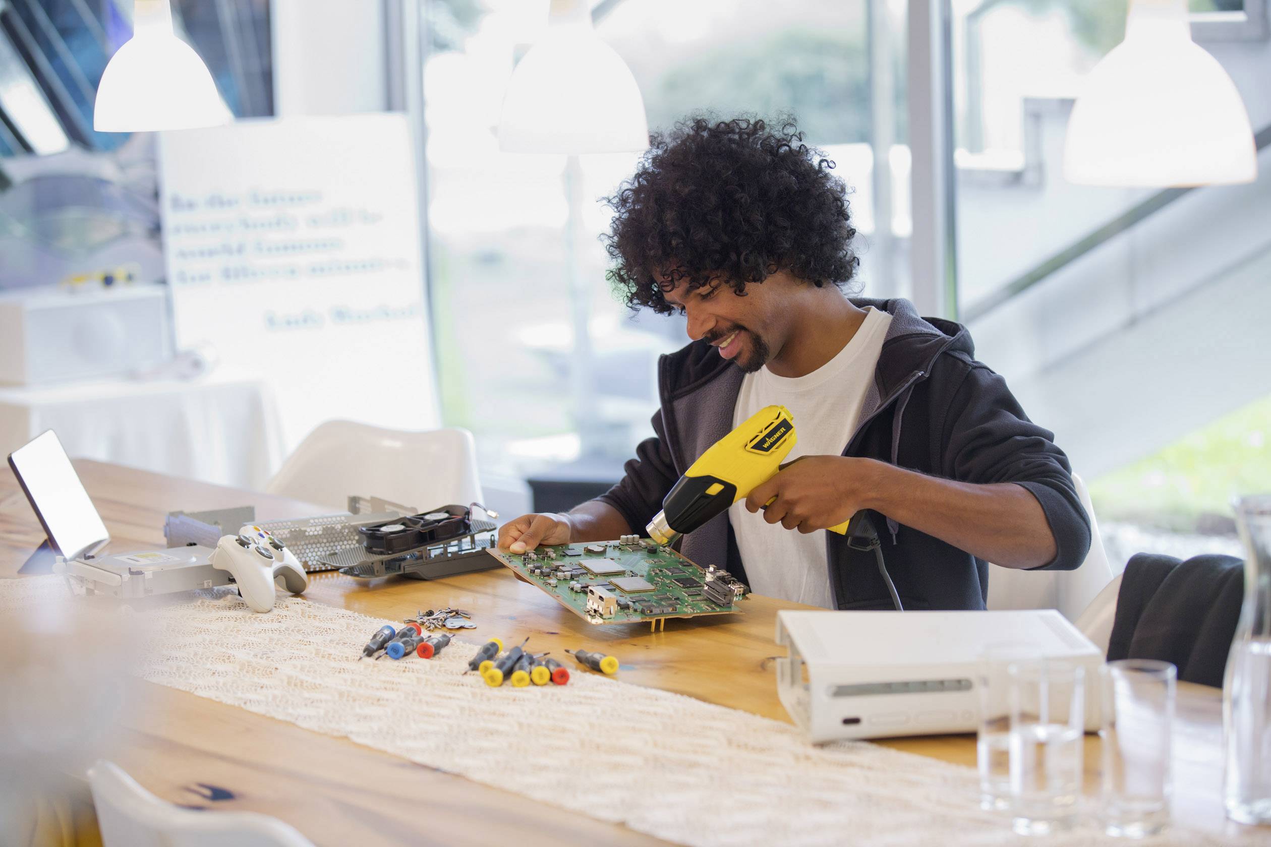 A man with curly hair is sitting at a table and repairing a circuit board with a tool. Electronic devices and tools are scattered across the table.