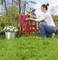 A woman is removing paint from a red chair with an electric tool in the garden. Flowers and a wooden fence are in the background.