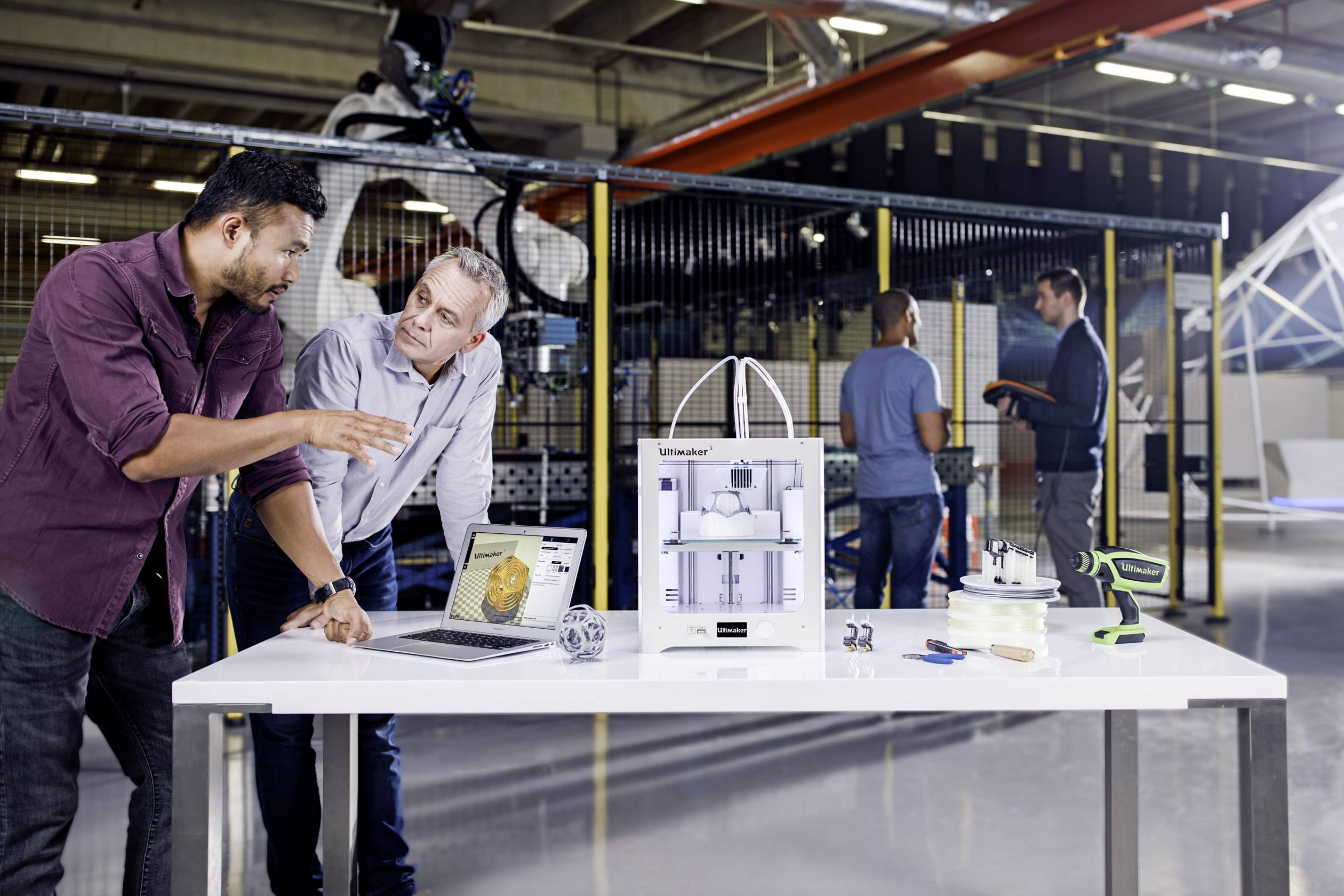 Two men are discussing a 3D printer on a table in a workshop. Other people are working in the background.