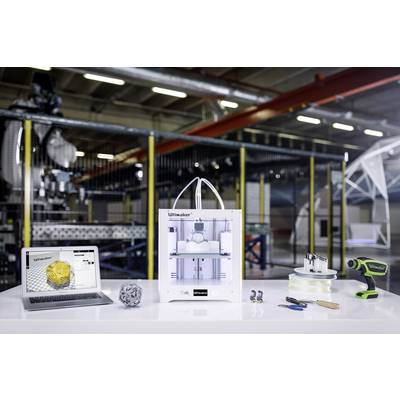 A 3D printer is positioned on a table, surrounded by a laptop, tools, and filament reels in an industrial setting.