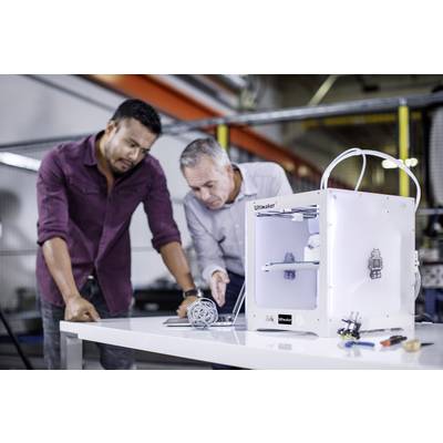 Two men are standing next to a 3D printer and examining a printed model. The printer is situated on a table in a workshop.