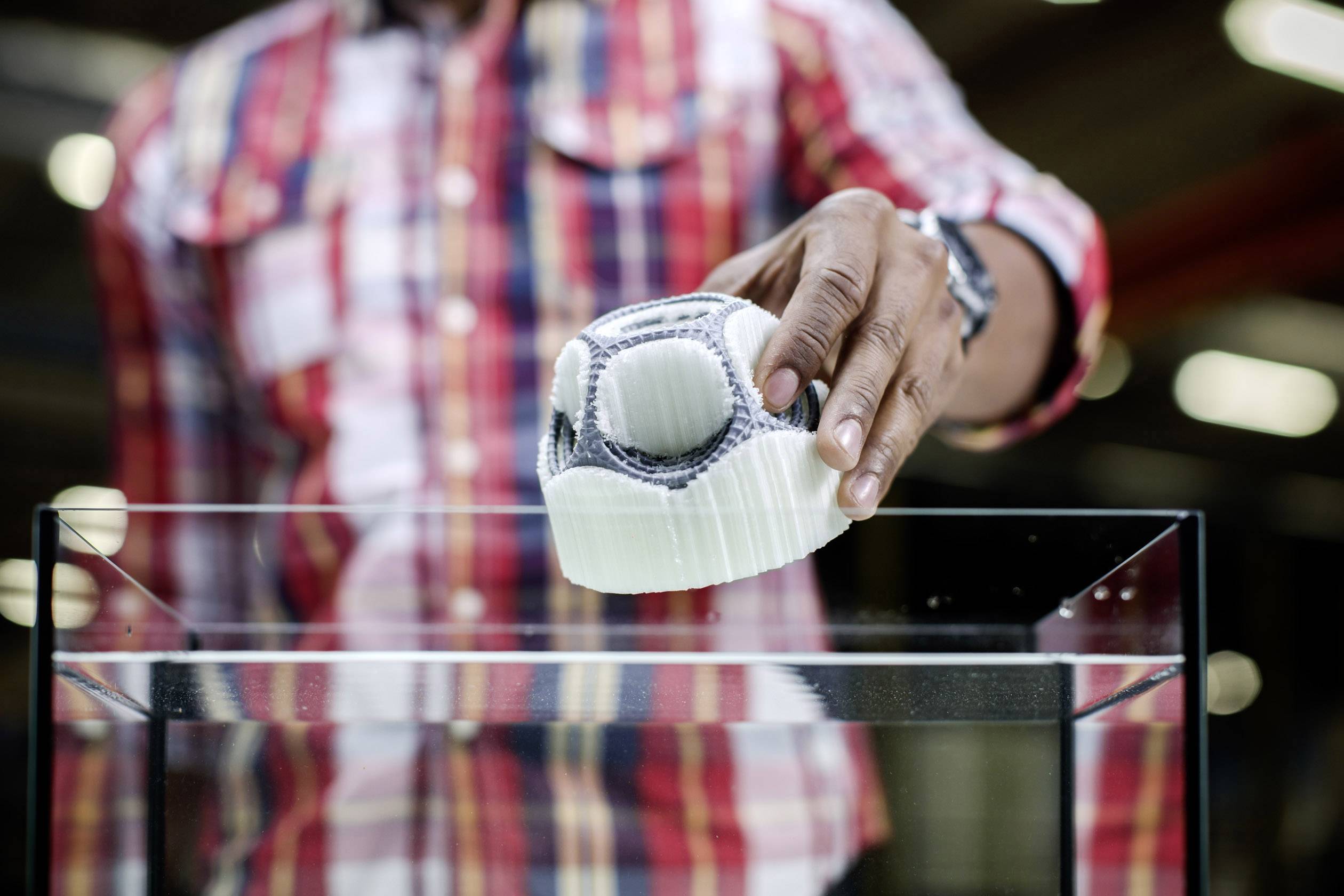 A person places a white 3D-printed shoe on a glass display cabinet. A checked shirt is visible in the background.