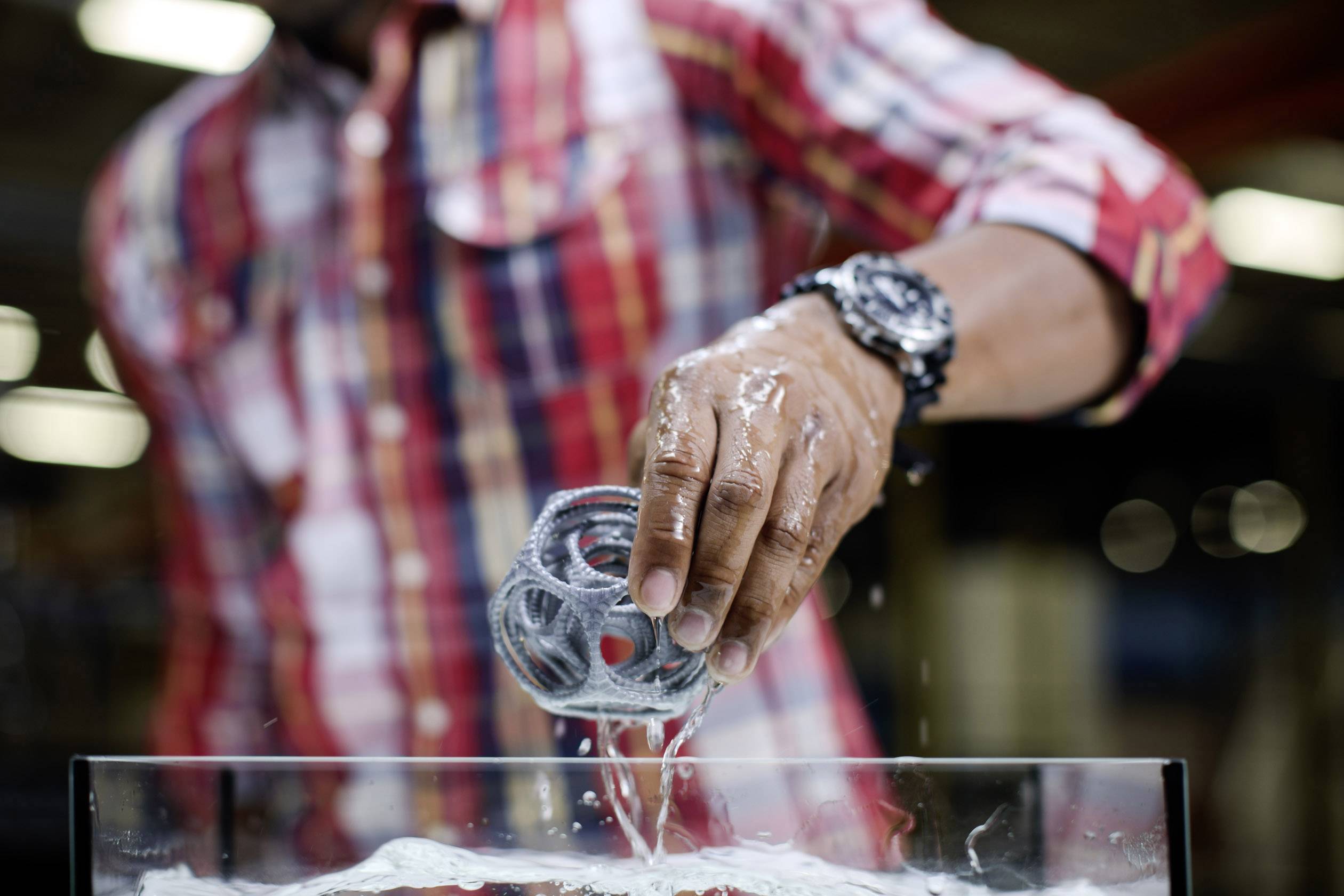 A person in a checked shirt is holding a wet metal object over a glass container filled with water.
