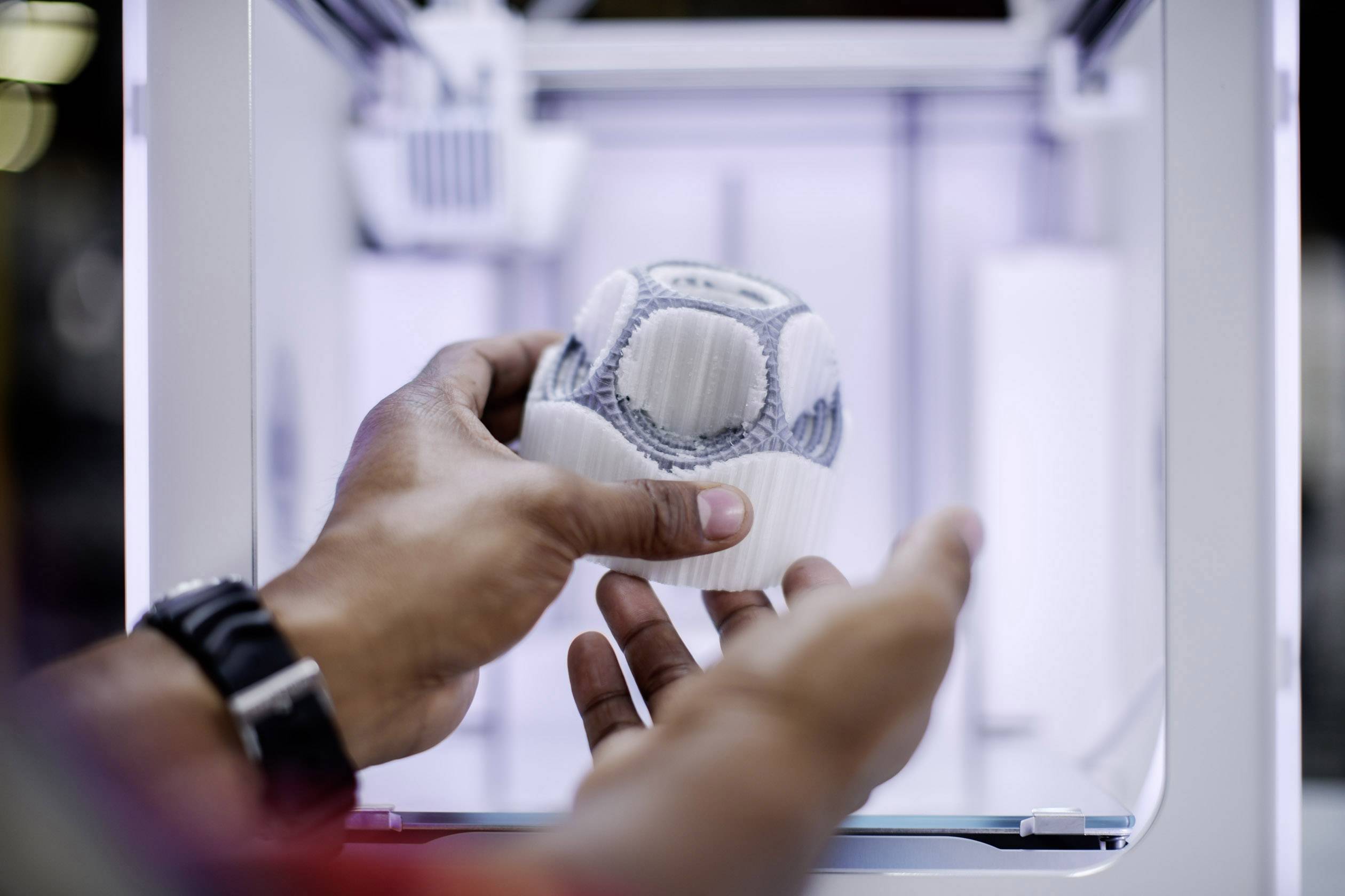 Hands are holding a 3D-printed, spherical object in a modern 3D printer.