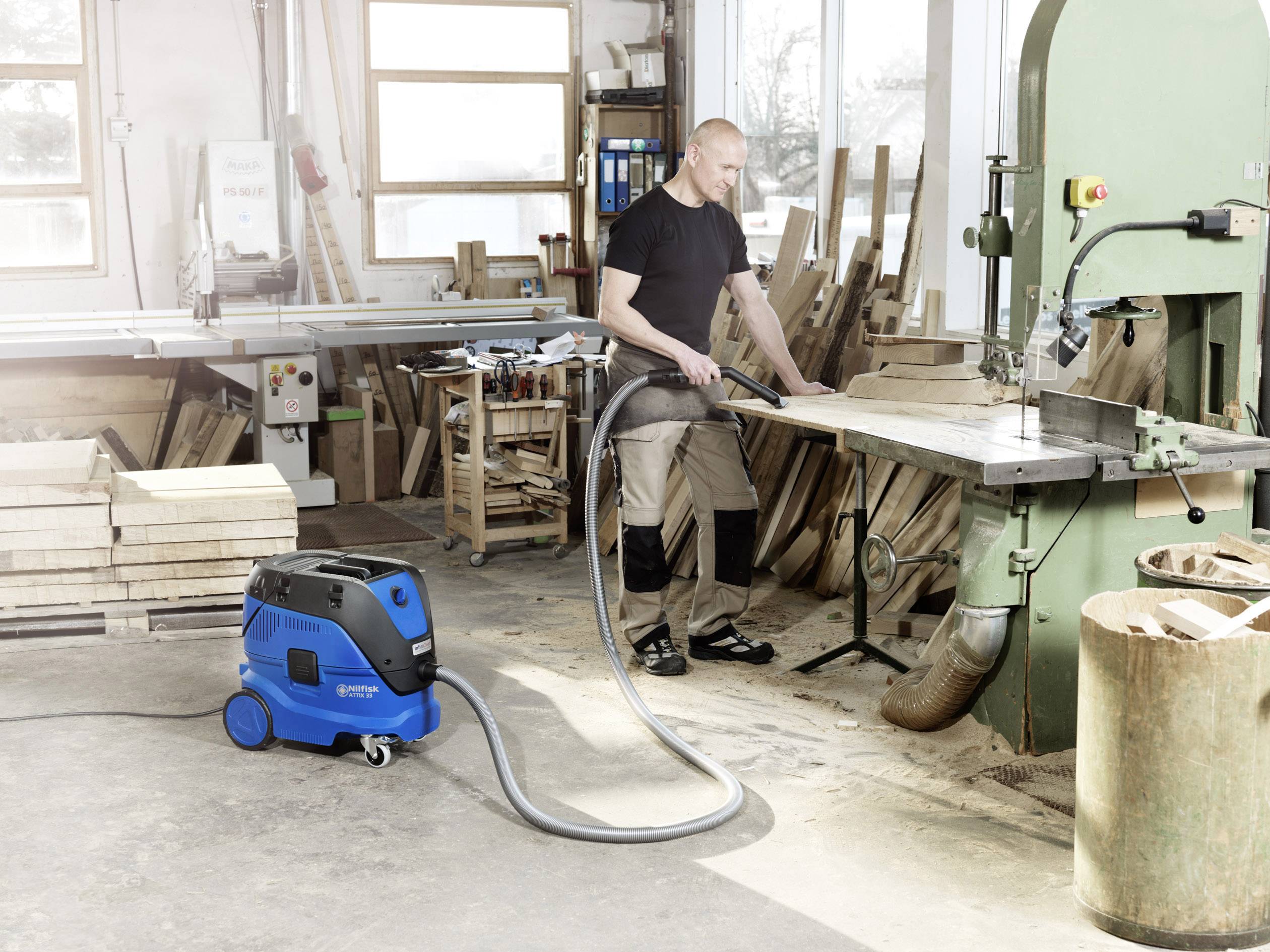 A man in a workshop is working with a bandsaw. The floor is clean, as a blue industrial vacuum cleaner is extracting the wood dust.