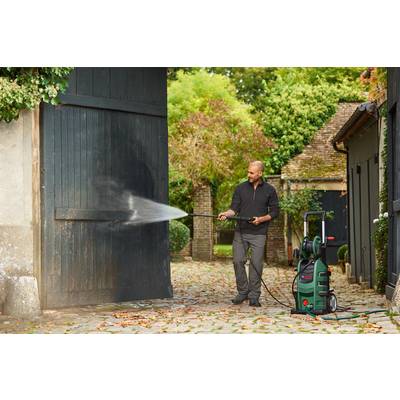 A man is cleaning a wooden gate with a pressure washer in a garden. Surrounded by foliage and trees in daylight.