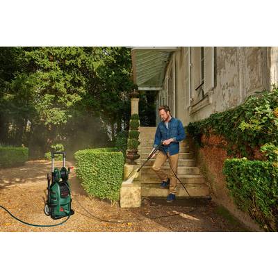 A man is cleaning the steps in front of a house with a pressure washer, surrounded by garden plants.