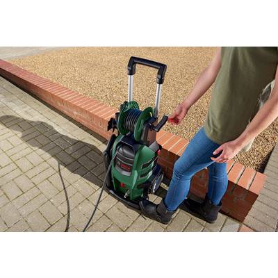 A person is operating a pressure washer with a green casing and a coiled hose, standing on a paved surface next to a wall.
