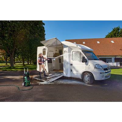 A person is cleaning a white motorhome with a pressure washer on a sunny car park in front of a building with a red roof.