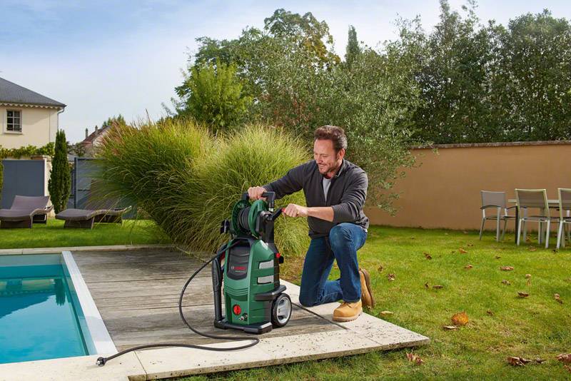 A man is kneeling by the swimming pool and cleaning its surface with a pressure washer. Garden furniture and plants can be seen in the background.
