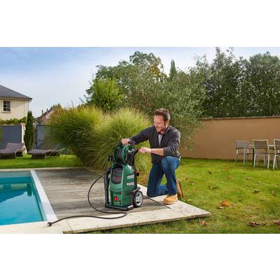 A man is kneeling by the swimming pool and cleaning its surface with a pressure washer. Garden furniture and plants can be seen in the background.