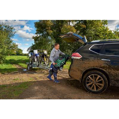 A man is loading a rucksack into the boot of an SUV. In the background, two motorbikes are on a trailer in a rural setting.