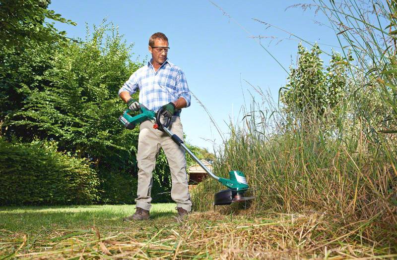 A person wearing safety glasses and gloves is using a lawn trimmer to cut long grass in a garden area.