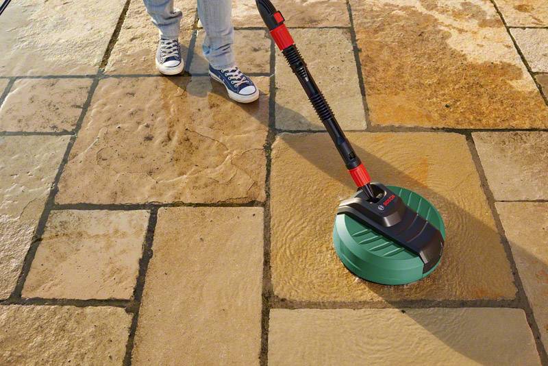 A person is cleaning a stone tile floor with a green high-pressure washer. The distinctive cleaning effect is clearly visible.