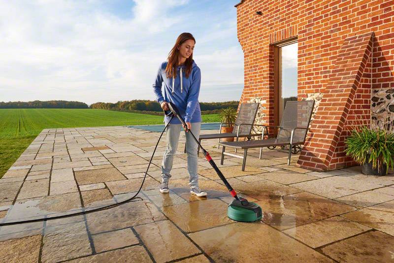 A woman is cleaning a patio with a pressure washer. In the background, there is a brick house and a spacious green lawn.