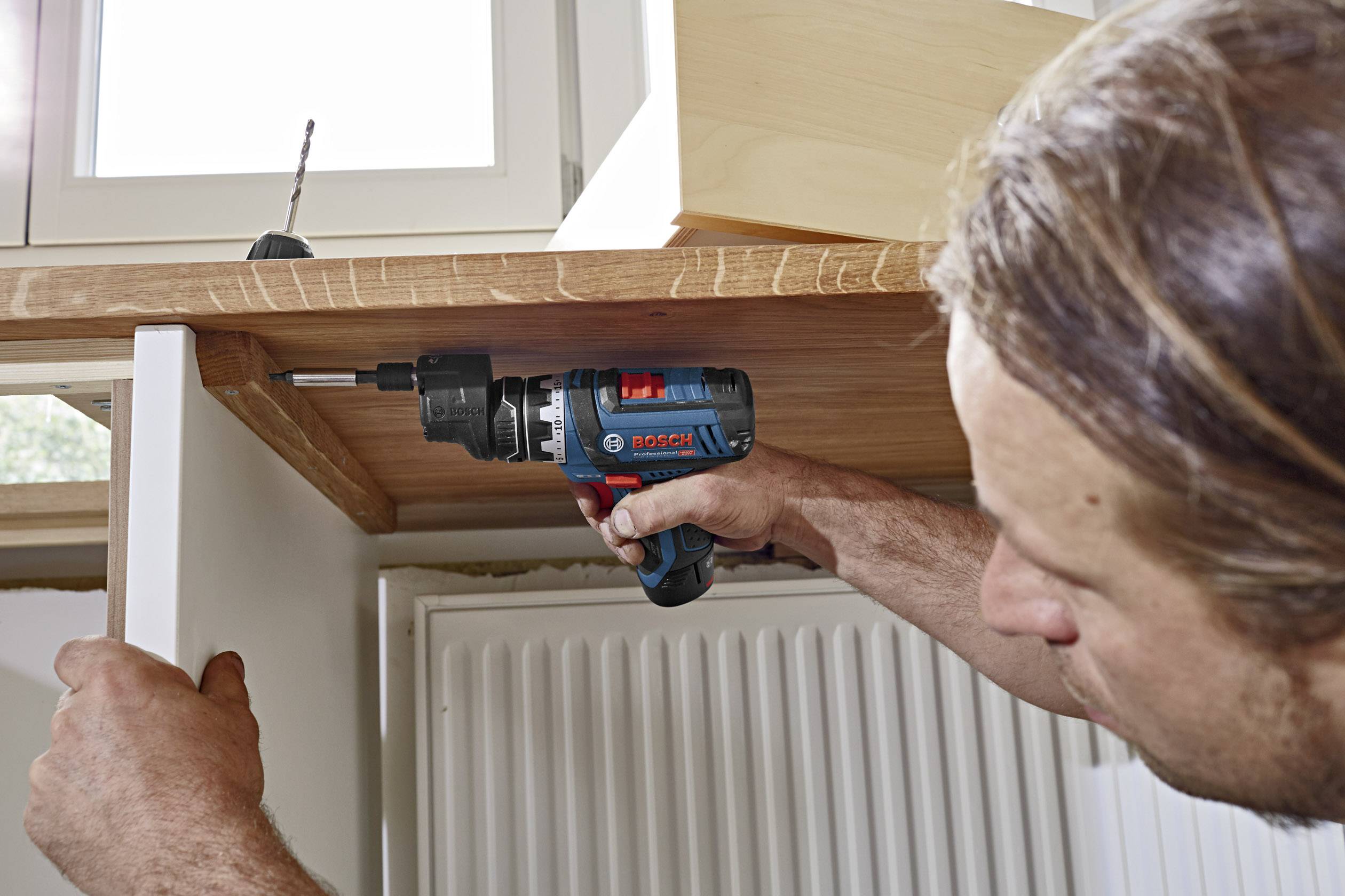 'A man is installing a shelf board with an electric drill beneath a window. A radiator is visible in the background.'