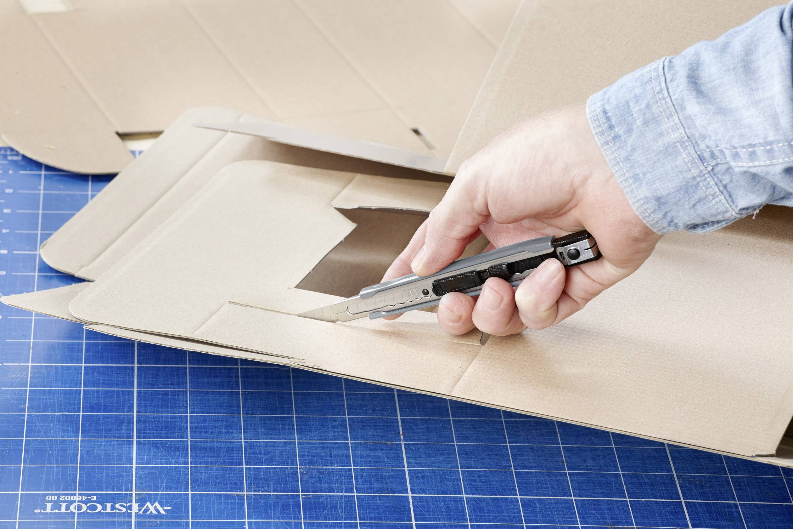 A hand is using a craft knife to cut a flat cardboard box on a blue cutting mat with a grid pattern.
