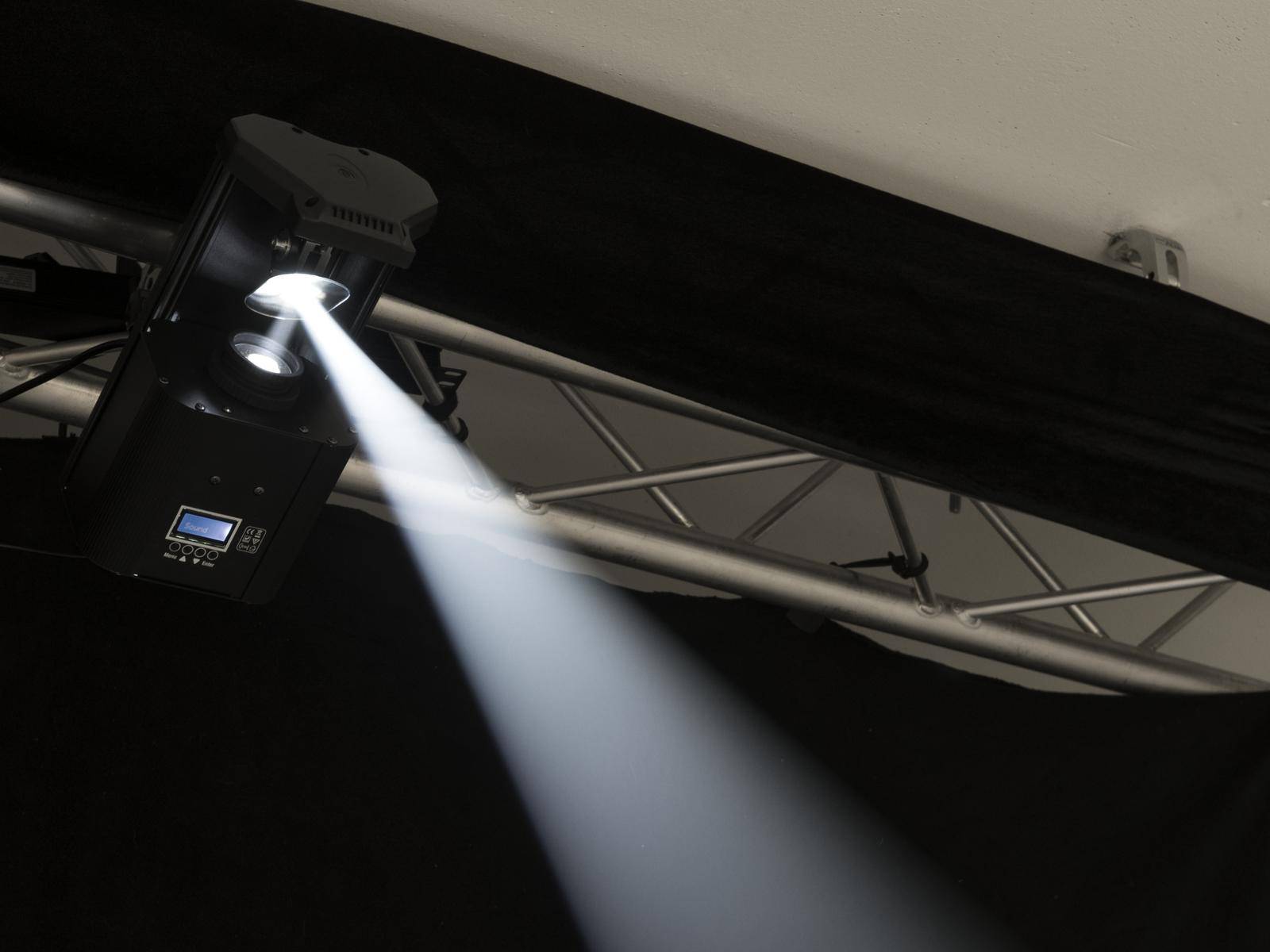 A black spotlight hangs from a metal frame and projects a beam of light in a dark room.